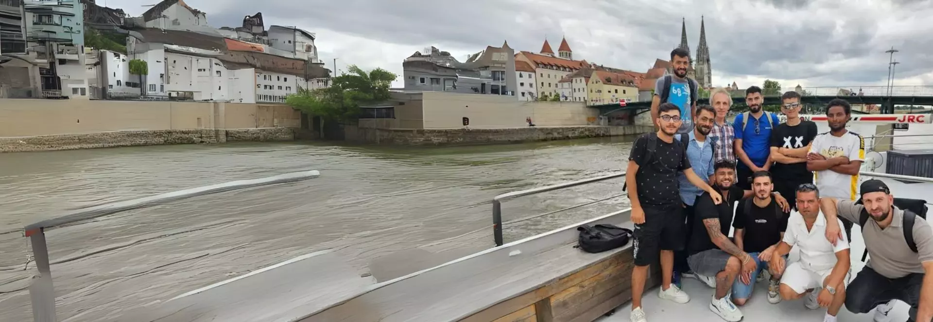 Auf dem Bild sieht man rechts eine Gruppe von Männern, welche sich auf einem Schiff befinden. Im Hintergrund sieht man die Donau fließen sowie eine Kulisse von Regensburg