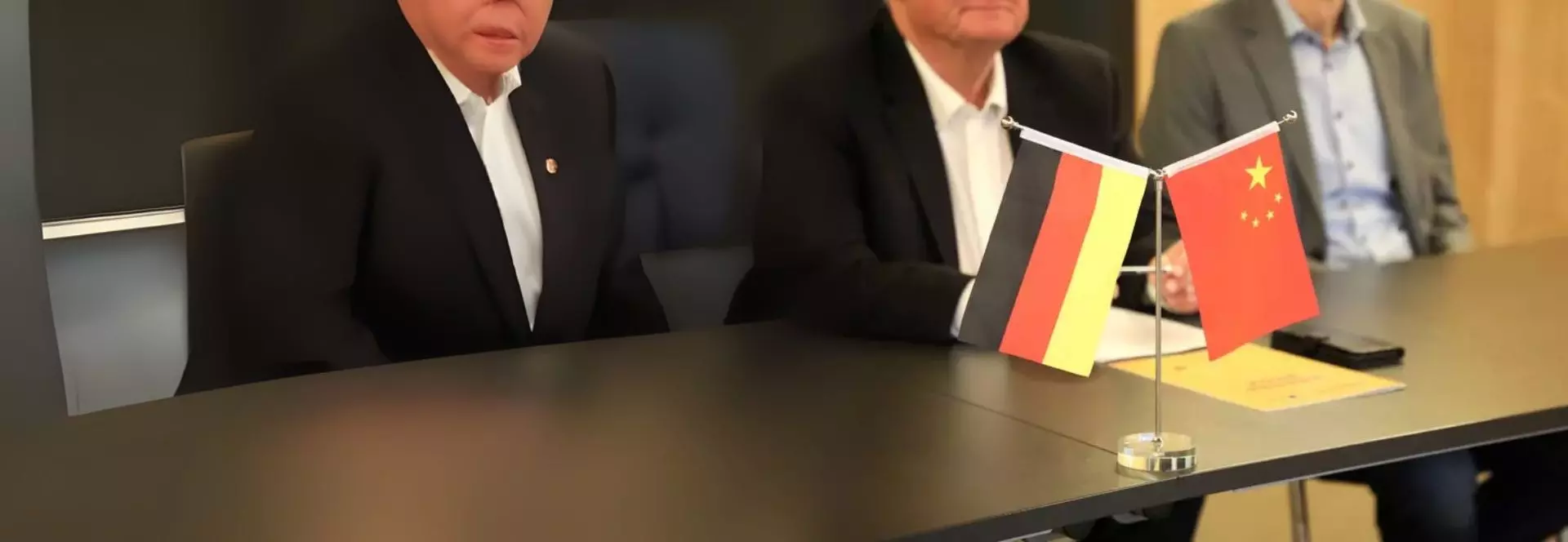Auf dem Bild sind drei Personen zu erkennen, welche an einem Tisch sitzen. Die Gesichter sind nicht erkennbar. Auf dem Tisch steht eine kleine Doppelfahne mit der Deutschland- und der Chinaflagge.